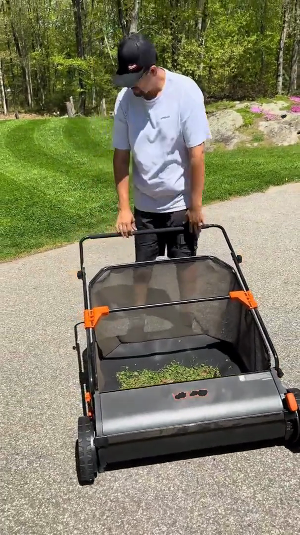 A push lawn sweeper moving down a driveway with a hopper full of clippings